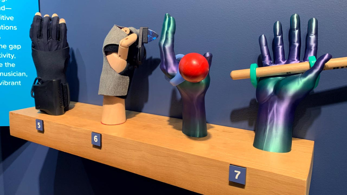 Adaptive and prosthetic hands gripping objects, including a red ball and a wooden stick, on a numbered museum display shelf