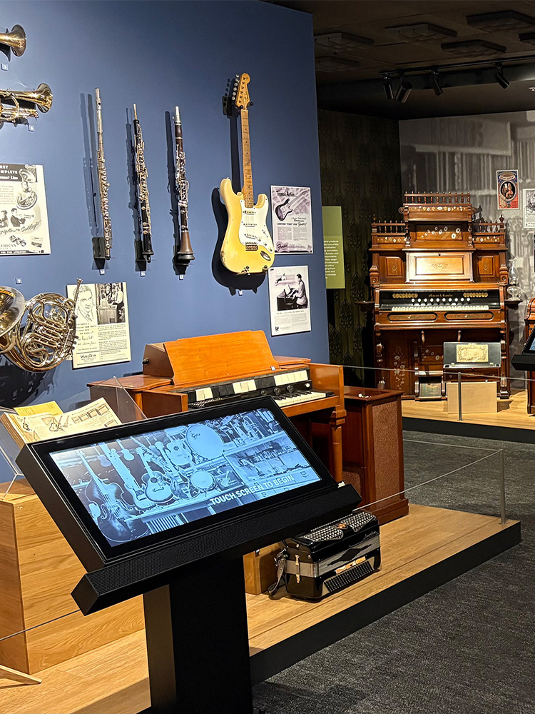 Brass instruments, a guitar, and a vintage organ on display with a touchscreen kiosk at the Museum of Making Music