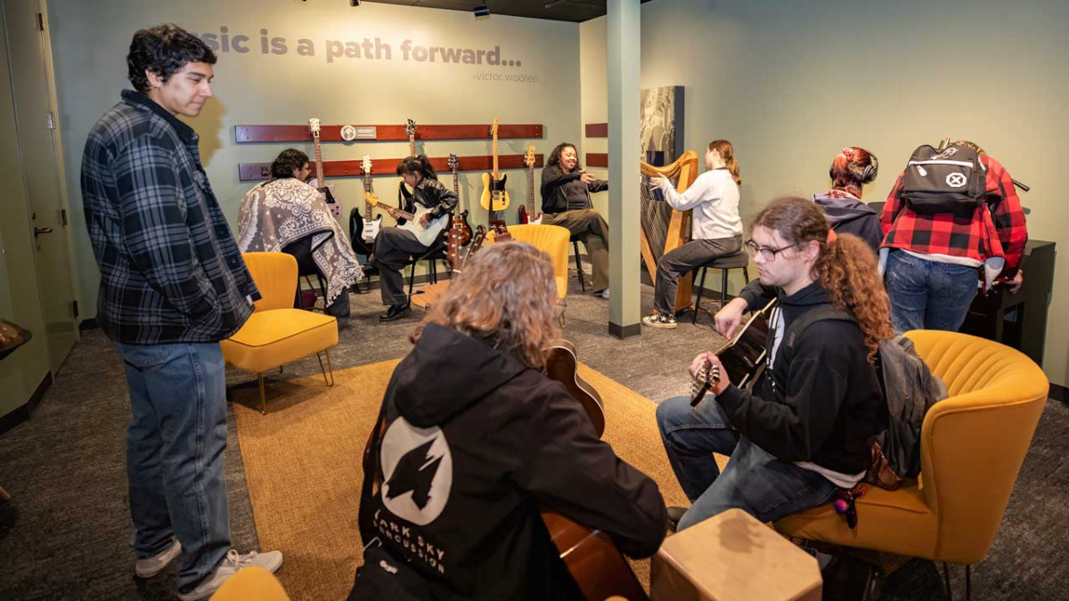 Students play guitars, a harp, and a cajón in a museum room beneath a wall of hanging guitars