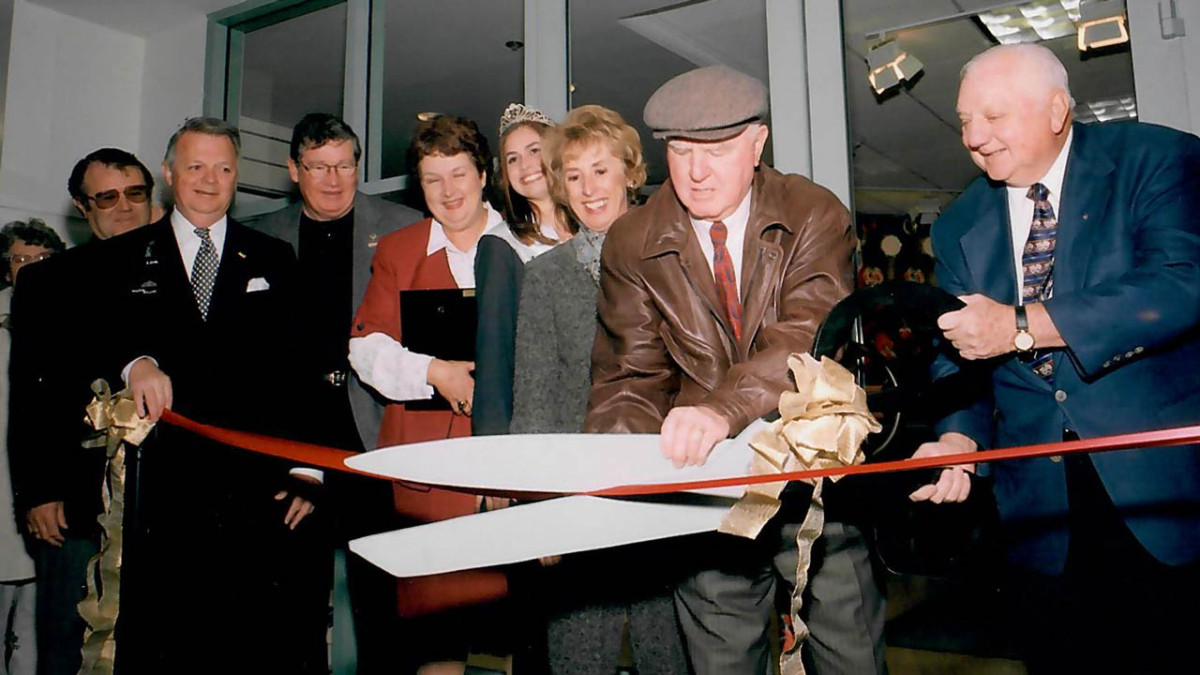 Archival photo of a group cutting a red ribbon with oversized scissors at a museum ribbon-cutting ceremony