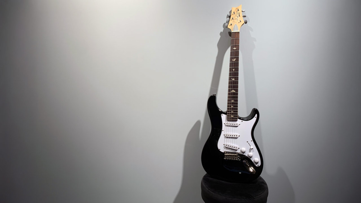 A black PRS electric guitar with a white pickguard and bird fret inlays leaning on a stool against a gray wall