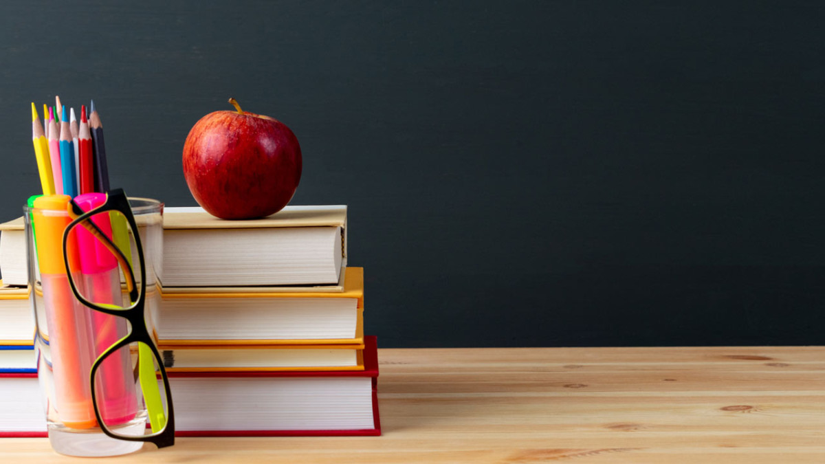 Stacked books topped with a red apple beside a cup of colored pencils and eyeglasses in front of a chalkboard