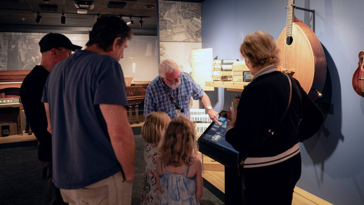 A volunteer shows a family an interactive touchscreen in a gallery of keyboards and mandolins