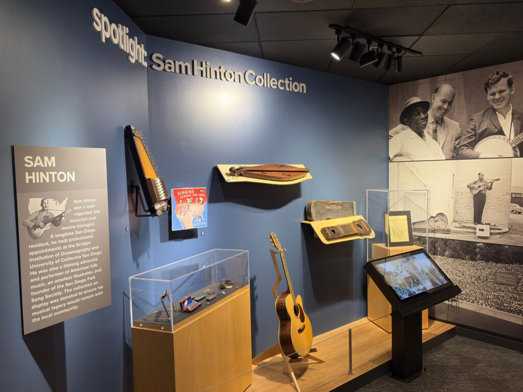 Sam Hinton Collection gallery with an introductory text panel, wall-mounted folk instruments, display cases, an acoustic guitar, and a touchscreen kiosk
