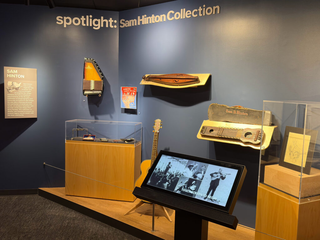 Wide view of the Sam Hinton Collection with folk instruments mounted on a blue wall above wooden display cases