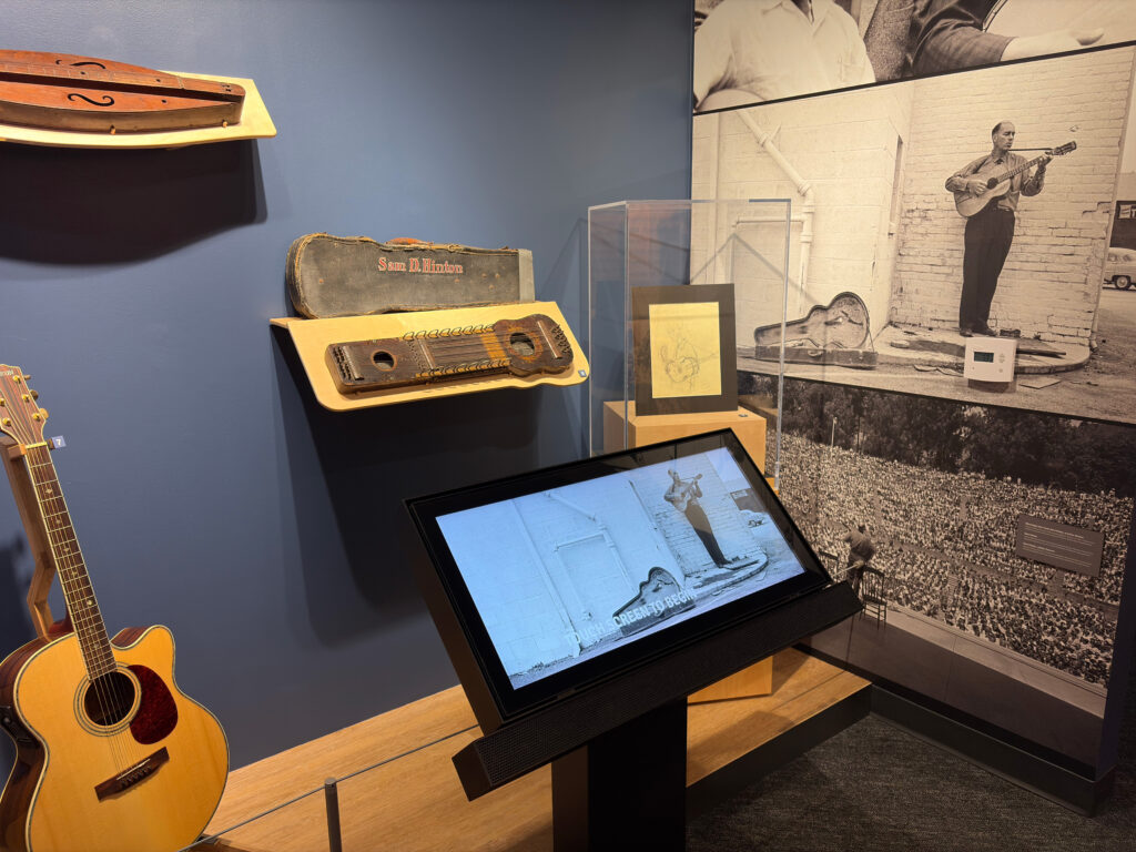 Acoustic guitar and touchscreen kiosk in front of a large black-and-white photo of Sam Hinton performing
