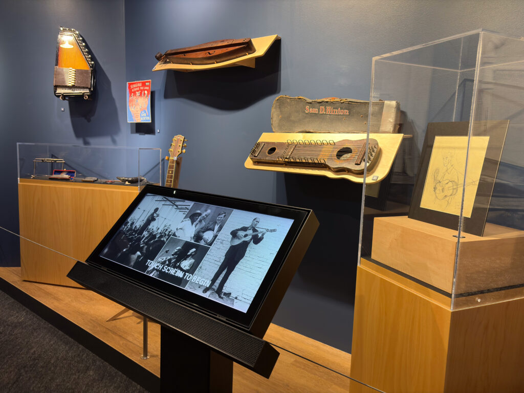 Touchscreen kiosk showing archival photos in front of wall-mounted folk instruments and display cases