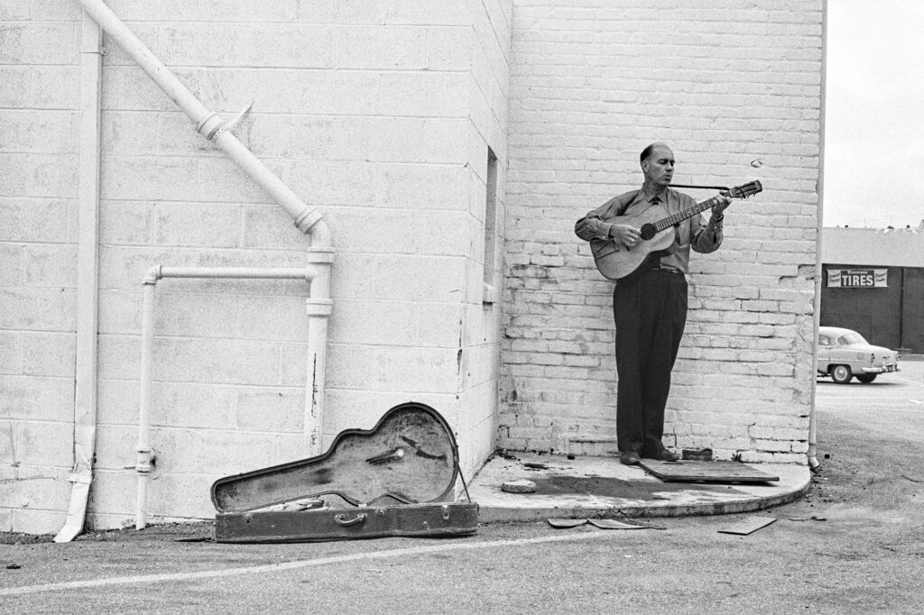 Sam Hinton playing guitar against a white brick wall with an open guitar case on the ground beside him
