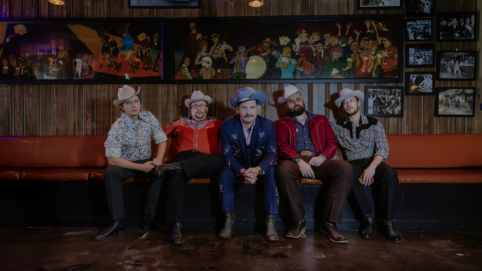 The Po' Ramblin' Boys, five band members in cowboy hats and embroidered Western shirts, seated on a leather banquette beneath a painted mural and framed photos