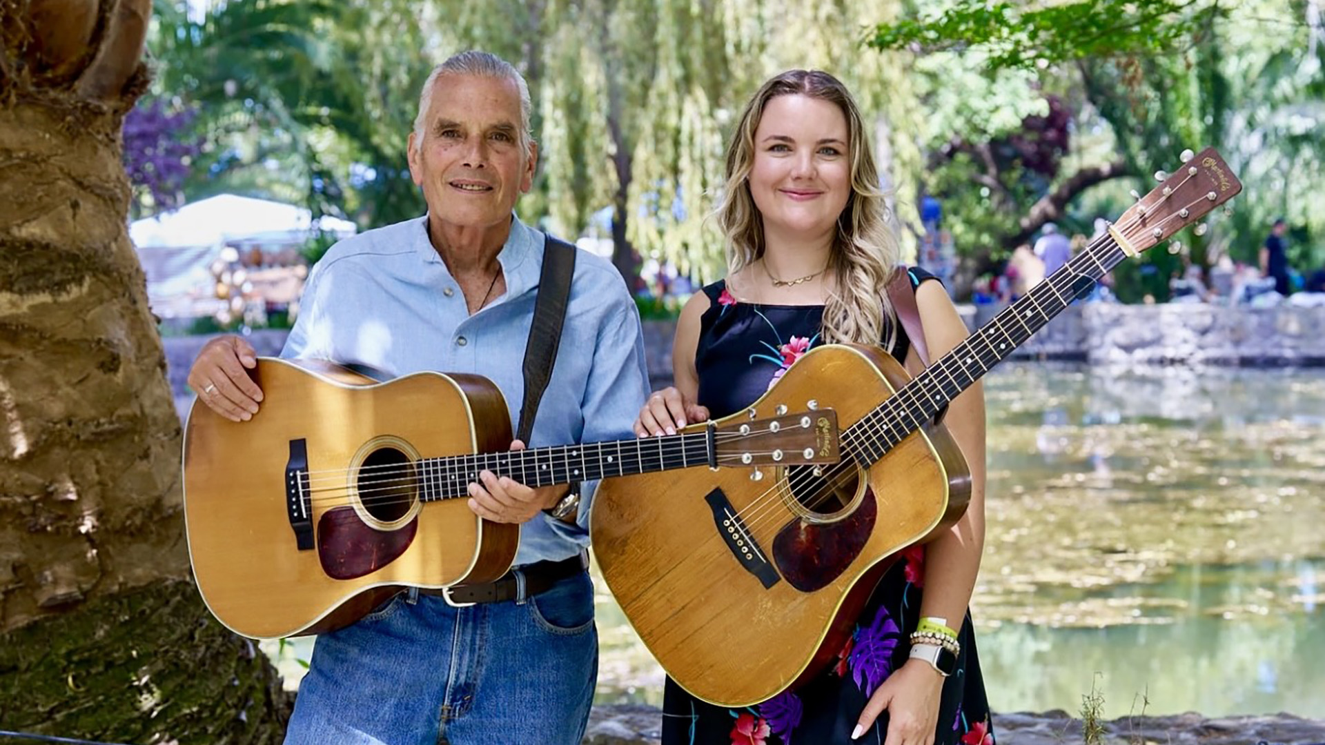 Keith Little and Joy Louise holding acoustic guitars outdoors by a pond